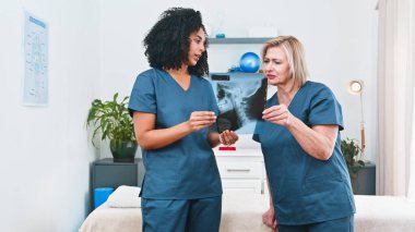 Two healthcare professionals in blue scrubs examine an X-ray in a clinical room, discussing diagnosis and next steps for rehabilitation. Calm, collaborative setting with medical equipment and plants in the background.