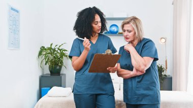 Two female healthcare professionals in blue scrubs review a patients plan using a clipboard in a bright physiotherapy clinic. They discuss exercises, progress, and care, showing professional support and teamwork.