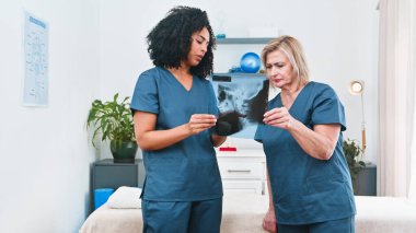 Two physiotherapists in blue scrubs examine an X-ray in a treatment room, discussing care plans and sports therapy. The professional, collaborative atmosphere supports patient rehab and wellness.