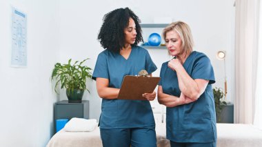 Two healthcare professionals in blue scrubs discuss a treatment plan with a senior patient in a clinic setting, holding a clipboard. Compassionate, professional care and guidance in physiotherapy.