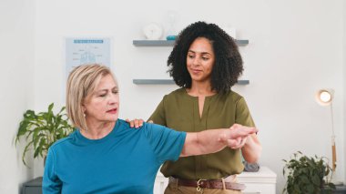 A friendly physiotherapist assists an elderly woman with a gentle arm stretch in a clinical therapy room. Warm lighting, plants and charts create a calm, professional setting focused on rehabilitation and well-being.