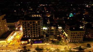 Hava karardıktan sonra hareketli bir kıyı şehrinin nefes kesici yüksek açılı perspektifi. Parlak şehir ışıkları otelleri, sokakları ve gece hayatını aydınlatıyor.