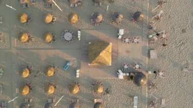 Zenithal hava perspektifi, temiz düzenlenmiş şemsiyeleri ve şezlongları olan ıssız kumlu bir sahilde gün batımında uzun gölgeler bırakarak uçuyor.