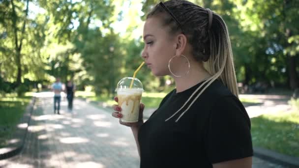 Jovem bela trançado fêmea beber milkshake com caramelo no parque ensolarado — Vídeo de Stock