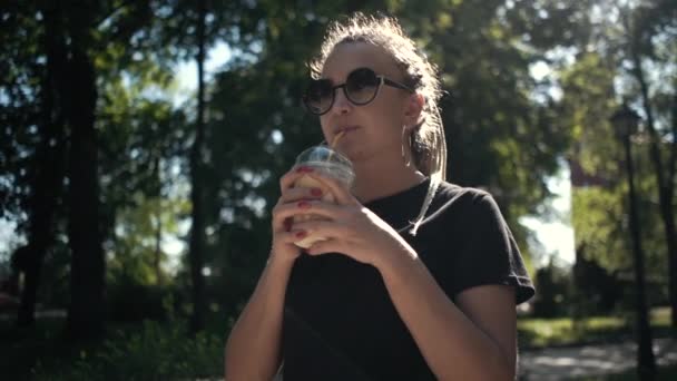 Молодая красивая плетеная женщина пьет молочный коктейль с карамелью в солнечном парке — стоковое видео