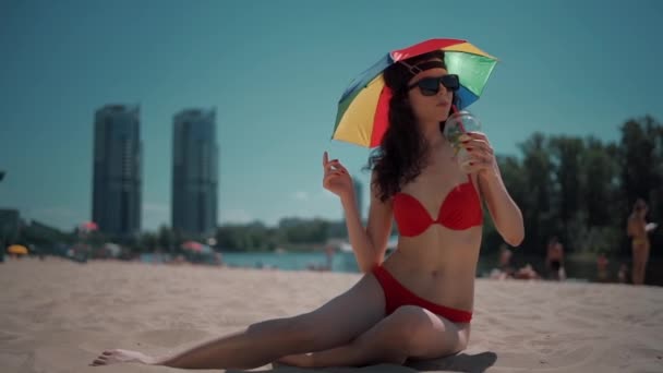 美しい女性は傘を持って街のビーチで休憩しモヒートカクテルを飲む — ストック動画