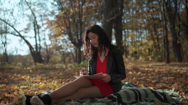 Atraente encaracolado morena mulher sentado e escrevendo no bloco de notas no parque no outono — Vídeo de Stock