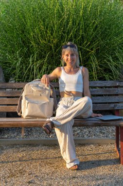 Hafif giysili gülümseyen genç bir kadın bankta oturuyor ve kameraya bakıyor. Rahatlamış ruh hali, olumlu duygular, doğal ışık. Açık hava yaşam tarzı, güven kavramı.