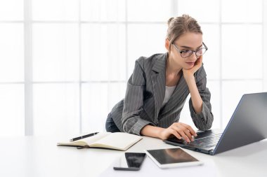 Güzel bir kadın evde dizüstü bilgisayarıyla çalışıyor. Kadın gözlük takıyor ve gri takım giyiyor..