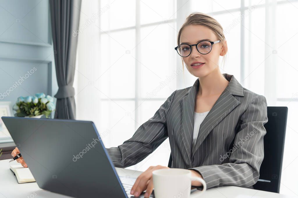 Beautiful woman is working at home with her laptop. The woman is wear glasses and grey suit.