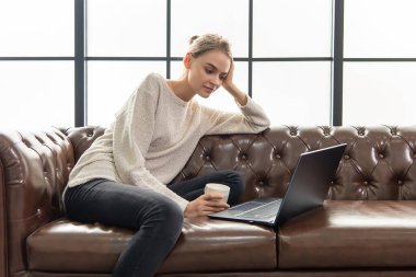 Zeki iş görünüşlü güzel bir kadın deri koltukta oturuyor ve elinde kahve fincanı var. Evden çalışıyorum. Sıcak oturma odası.