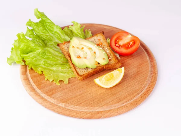 Wooden plate with toasted bread, slices of avocado and tomato on white background