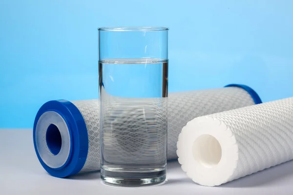 A glass of clear water and filter cartridges to domestic water treatment systems at blue background