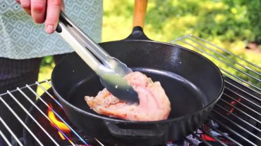 Bir parça marine edilmiş tavuk fileto dışarıda ızgarada kızartılıyor. Aşçı ızgaraya bir parça tavuk filetosu koymak için metal maşa kullanır. Arka bahçede et kızartıyoruz..