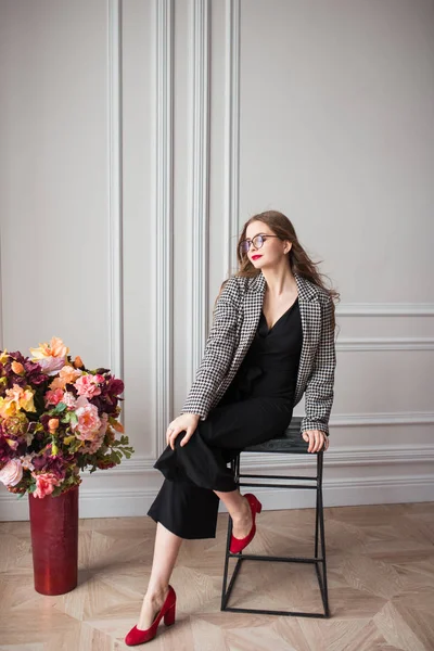 young beautiful fashion model in black dress and stylish blazer and glasses sitting in chair with flowers indoors