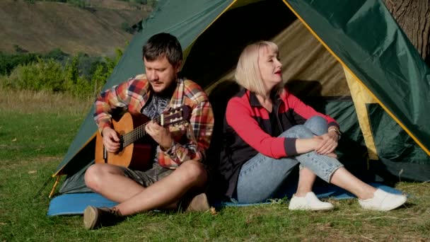 Uomo e donna cantano una canzone con la chitarra seduta alla tenda. La famiglia è in campeggio. — Video Stock