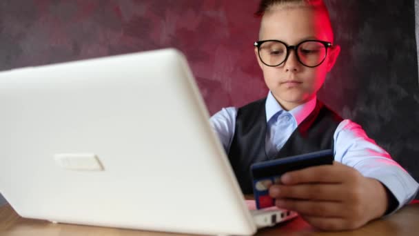 Modern child makes purchases on Internet with a laptop. Young boy with glasses makes an online order using a credit card. — Stock Video