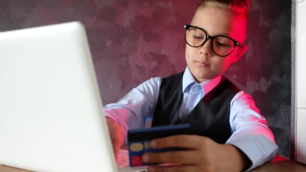 Niño moderno hace compras en Internet con un ordenador portátil. Niño con gafas hace un pedido en línea utilizando una tarjeta de crédito. — Vídeo de stock