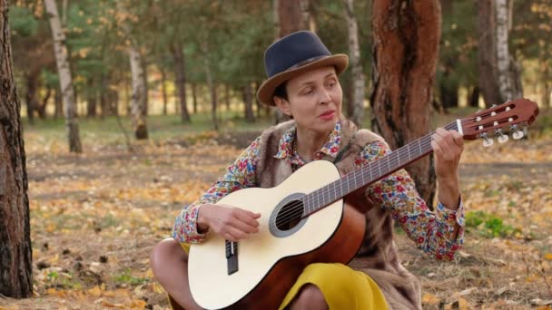 O femeie frumoasă într-o pălărie cântând la chitară acustică în parc. Un muzician de stradă care stă pe pământ cântă un cântec cerând bani . — Videoclip de stoc