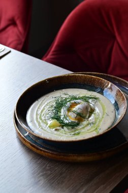 Dereotlu ve yumurtalı kremalı çorbaya yakın çekim. Bir restoranda, ahşap bir masada servis ediliyor. Orta Avrupa mutfağının sıcaklığını ve orijinalliğini yansıtan geleneksel ev yapımı bir yemek..