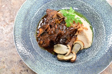 Soslu haşlanmış sığır yanaklarının üst görüntüsü, mantarlarla servis ediliyor ve mavi bir tabakta servis ediliyor. Geleneksel sonbahar yemeği gurme konforlu ev yapımı yemek olarak tasarlandı..