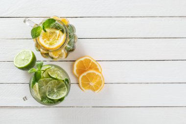 İki cam bardak kireç ve limon, ev yapımı limonata ile beyaz ahşap rustik zemin üzerine narenciye dilimlenmiş. Üstten görünüm. Kopya alanı