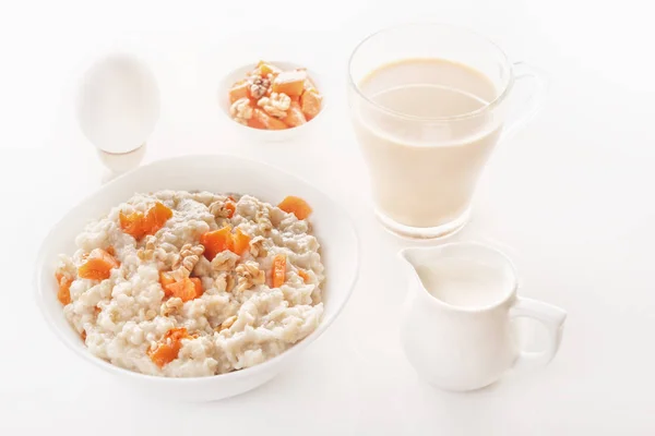 stock image Oatmeal with pumpkin and nuts in a plate, a glass of tea, a boiled egg and a jug with milk on a white background. Close-up.