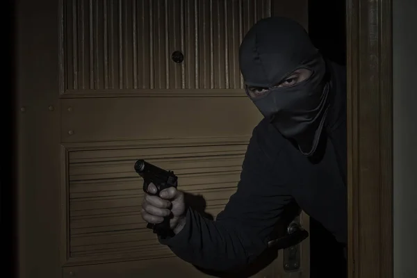 Ladro con una pistola in mano e un cappello con una fessura per gli occhi si intrufola in casa attraverso la porta di notte al chiaro di luna — Foto Stock