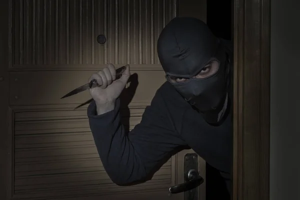 Ladrão com uma faca na mão e um chapéu com uma fenda para os olhos esgueiram-se para dentro da casa através da porta à noite ao luar — Fotografia de Stock