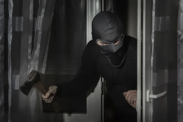 Ladro con un'ascia in mano e con un cappello con una fessura per gli occhi si intrufola in casa attraverso la porta di notte al chiaro di luna — Foto Stock