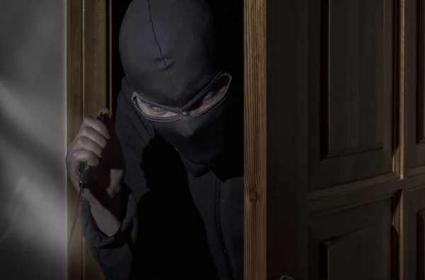 Thief with a knife in his hand and a hat with a slit for the eyes sneaks into the house through the door at night in the moonlight