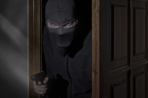 Ladro con una pistola in mano e un cappello con una fessura per gli occhi si intrufola in casa attraverso la porta di notte al chiaro di luna — Foto Stock