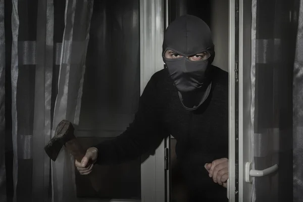 Ladro con un'ascia in mano e con un cappello con una fessura per gli occhi si intrufola in casa attraverso la porta di notte al chiaro di luna — Foto Stock