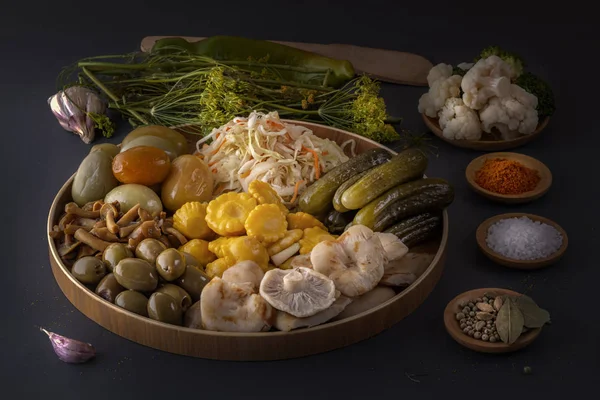 Alimentos fermentados repollo, pimientos, encurtidos, tomates, champiñones, calabacín, ajo en un gran plato de madera sobre una mesa oscura con especias para salar. Tecla baja . — Foto de Stock