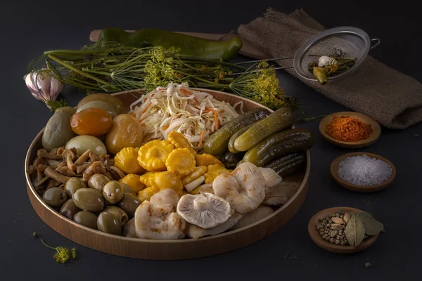 Fermented foods cabbage, peppers, pickles, tomatoes, mushrooms, zucchini, garlic in a large wooden dish on a dark table with spices for salting. Low key.