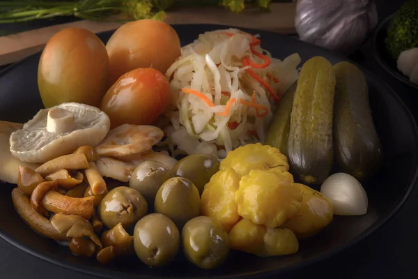 Alimentos fermentados repollo, pimientos, encurtidos, tomates, champiñones, calabacín, ajo en un plato negro en una mesa oscura — Foto de Stock