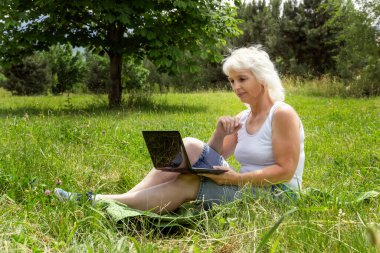 İnce, gri saçlı yaşlı bir kadın parkta güneşli bir çimenlikte oturur ve dizüstü bilgisayarla iletişim kurar.
