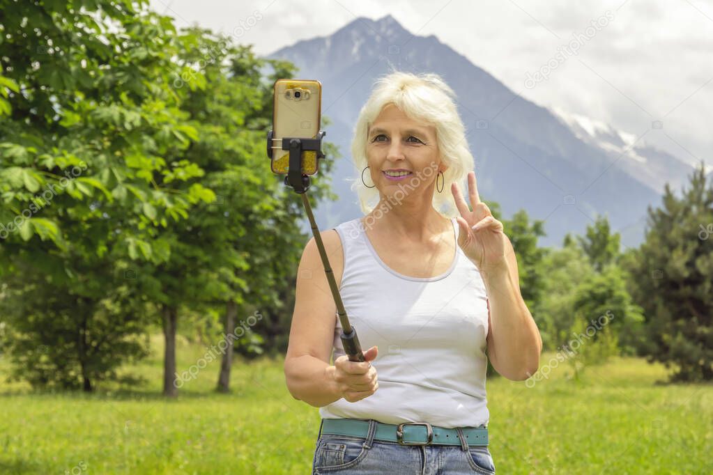 Slender elderly woman with gray hair takes a selfie and talks on a cell phone during a secluded walk in the park against the backdrop of the mountains
