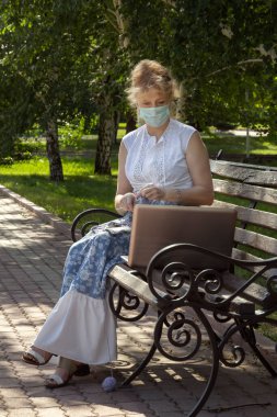 Yüzünde tıbbi maske olan bir kadın parkta oturmuş yün giysiler örüyor örgü örüyor ve ders izliyor ya da dizüstü bilgisayarla örgü örmeyi öğretiyor. Uzaklık öğrenimi