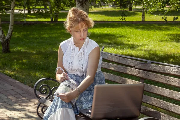 Woman in a park sits on a bench knitting wool clothes on knitting needles and watching lessons or teaching knitting through a laptop. Distance learning
