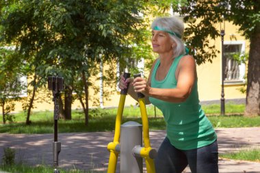 İnce, gri saçlı yaşlı bir kadın şehir parkındaki bir simülatörde bireysel fitness dersleri veriyor. Yakın plan.