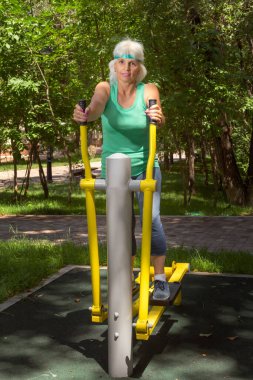 İnce, gri saçlı yaşlı bir kadın şehir parkındaki bir simülatörde bireysel fitness dersleri veriyor.