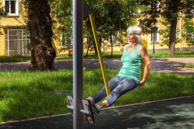 İnce, gri saçlı yaşlı bir kadın şehir parkındaki bir simülatörde bireysel fitness dersleri veriyor.