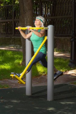 İnce, gri saçlı yaşlı bir kadın şehir parkındaki bir simülatörde bireysel fitness dersleri veriyor.