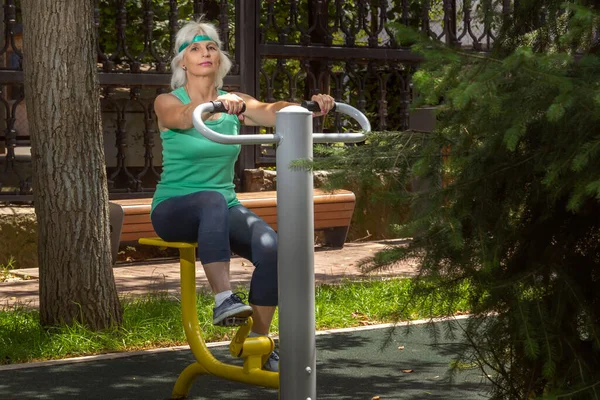 Slender elderly woman with gray hair conducts individual fitness classes on a simulator in a city park. Copy space