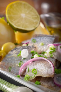 Kırmızı soğanlı, mayalanmış salatalıklı, patatesli, zeytinli ve limonlu ringa balığı dilimleri siyah ahşap bir masada beyaz bir tabakta. Yakın plan.