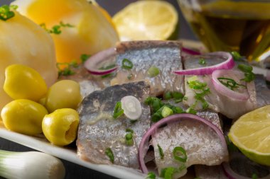 Kırmızı soğanlı, mayalanmış salatalıklı, patatesli, zeytinli ve limonlu ringa balığı dilimleri siyah ahşap bir masada beyaz bir tabakta. Yakın plan.