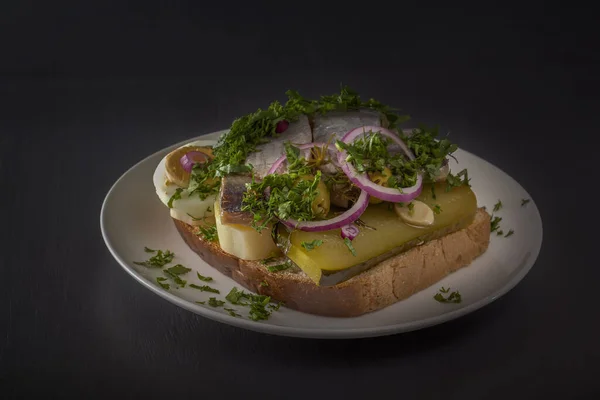 Sandwich with herring fillets, onions, pickled cucumber, egg and herbs on a white plate on a dark table. Traditional Scandinavian street food. Copy space