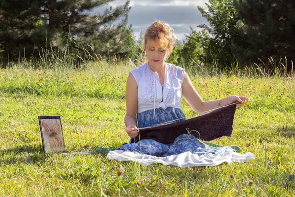 Woman sits on the grass in a park knitting wool clothes on knitting needles and watching lessons or teaching knitting through a laptop. Distance learning