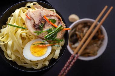 Siyah tabakta et ve yumurta parçaları olan erişte ve farklı geleneksel mantar, soya ve sebzelerle birlikte karanlık bir masa üzerinde loş meblağlar. Yukarıdan bak. Asya yemekleri konsepti. Yakın plan.